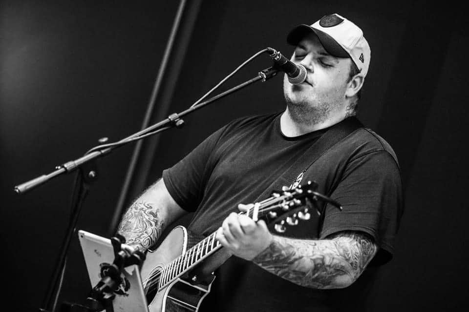 A man in a cap plays acoustic guitar and sings into a microphone with an electronic device on the stand. Black and white image.