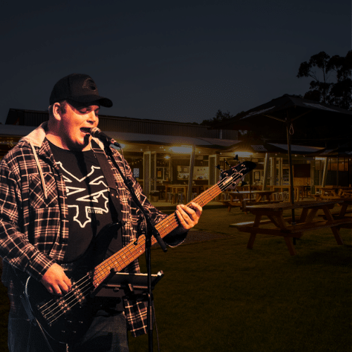 A man in a checked shirt and cap plays an electric bass guitar and sings into a microphone outdoors near picnic tables at dusk.