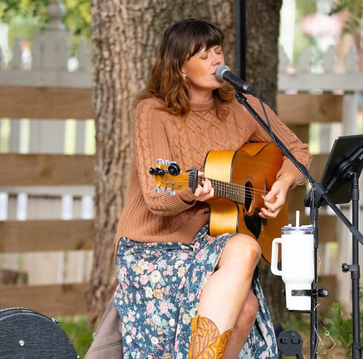Woman outdoors playing acoustic guitar and singing into a microphone; wearing brown jumper, floral skirt, mug nearby.