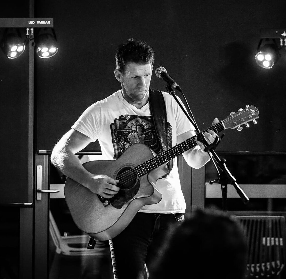 Man playing acoustic guitar and singing into microphone on stage under lights, wearing graphic T-shirt; black and white image.