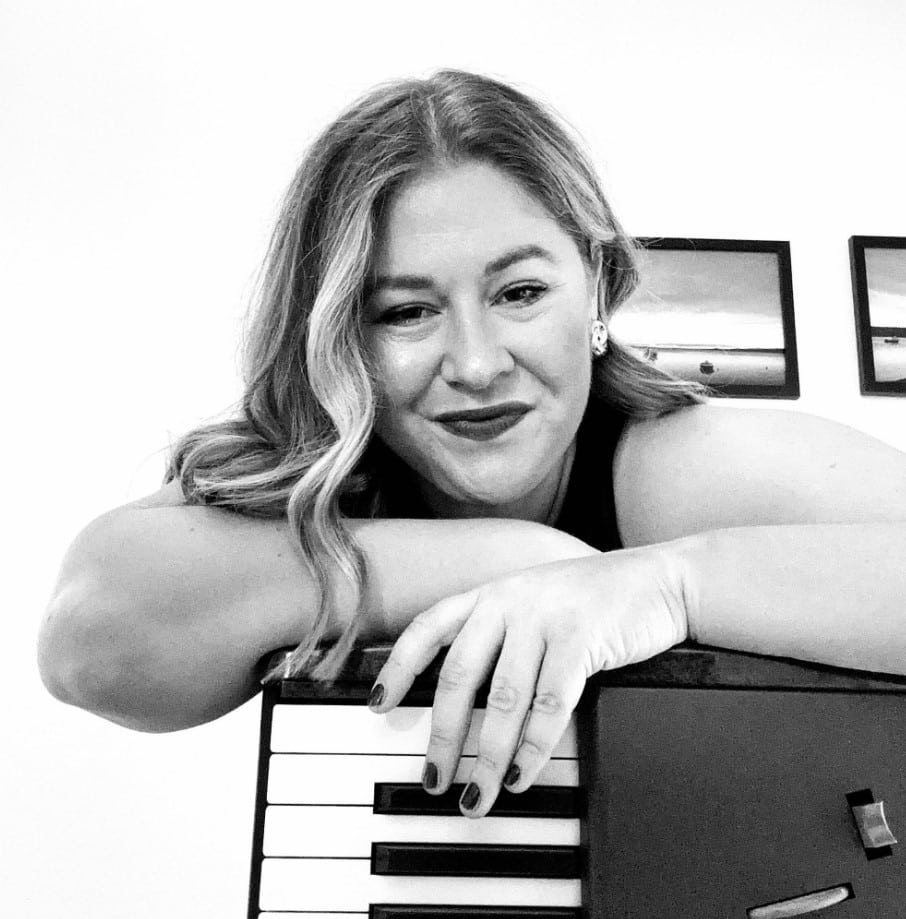A woman with wavy hair leans on a keyboard, smiling; framed pictures hang on the wall behind her.