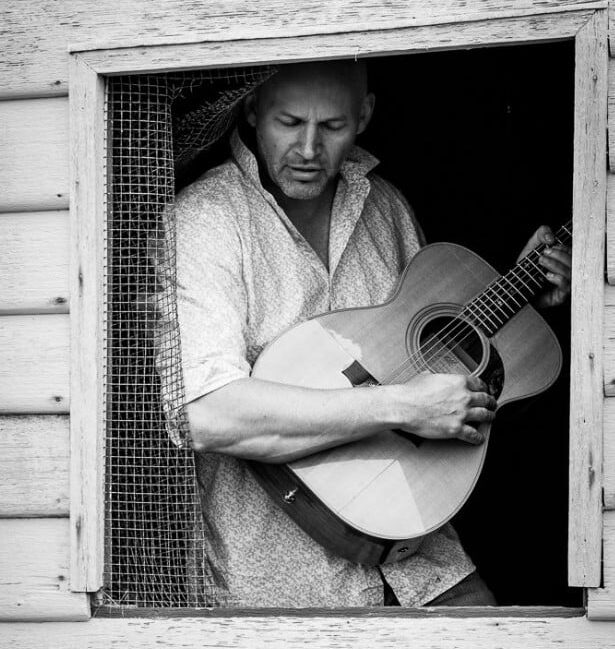 Man standing in a wooden building’s window frame, playing an acoustic guitar; black and white.