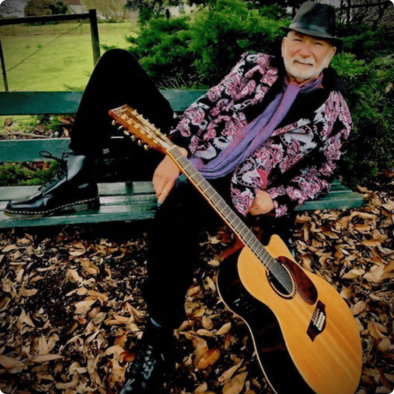 An older man in a patterned jacket and hat sits on a park bench holding an acoustic guitar, with fallen leaves and greenery around.