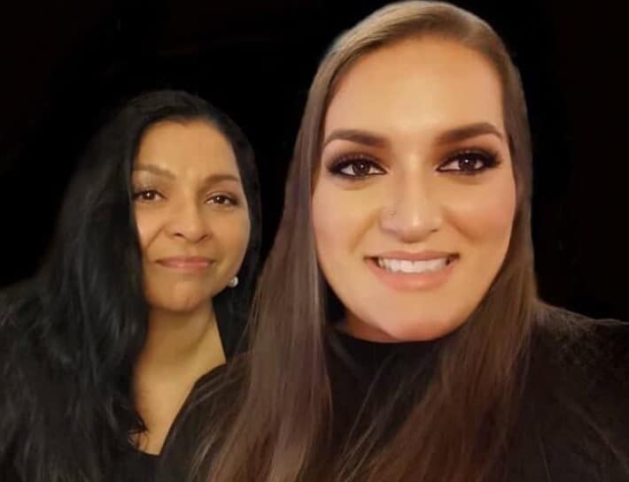Two women with long dark hair wearing black tops, smiling in front of a dark background.
