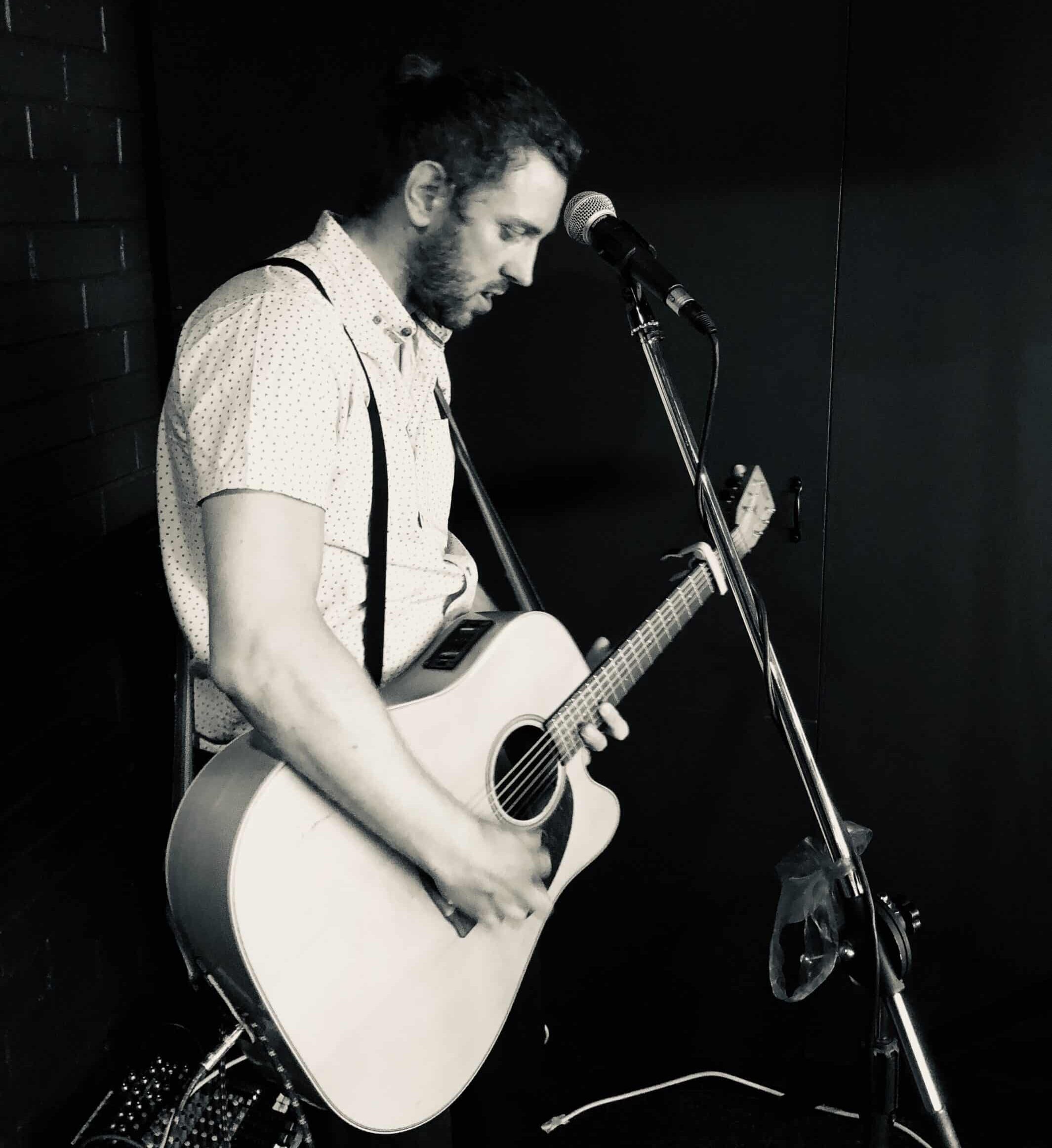 A man plays an acoustic guitar and sings into a microphone in a dimly lit room.