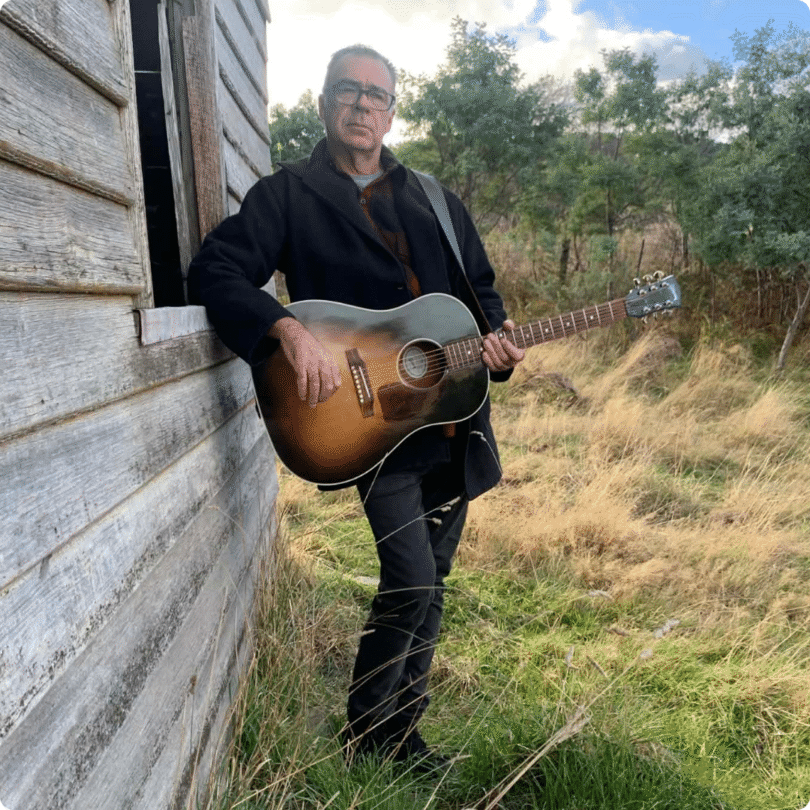 Man in dark coat holding acoustic guitar stands by weathered wooden building; trees and grass visible in background.