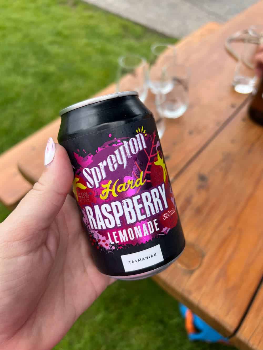 A hand holds a can labelled "Spreyton Hard Raspberry Lemonade" beside a wooden table with empty glasses outdoors.