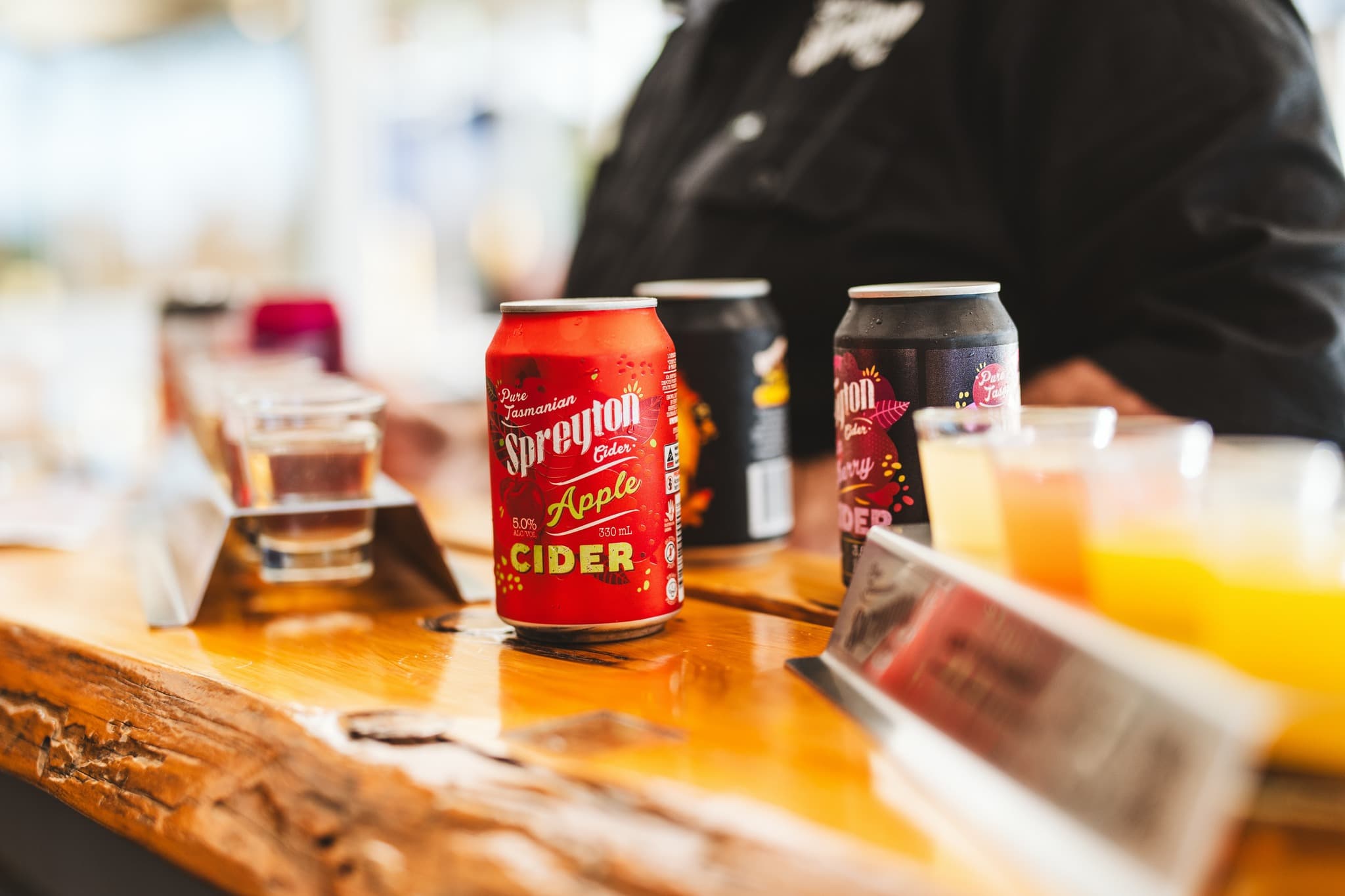 A red Spreyton Apple Cider tin on a wooden bar beside assorted drinks and beverage samples; a person is in the background.