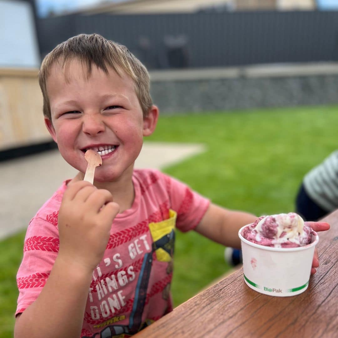 Boy in red shirt sits outdoors at a wooden table, holding a spoon and tub of ice cream, with grass in the background.