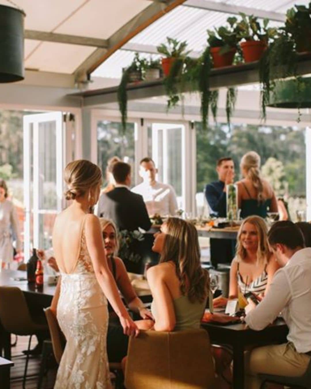 A woman in a white dress talks to guests seated indoors; sunlight, open doors, and potted plants are visible above.