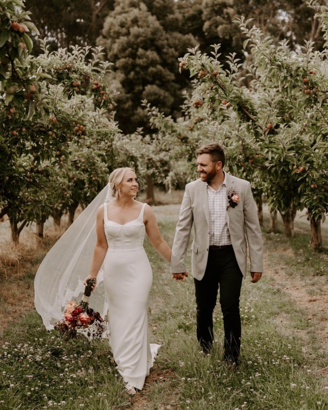 Bride and groom walk hand in hand through an orchard; the bride wears a long white dress, veil, and holds a bunch of flowers.