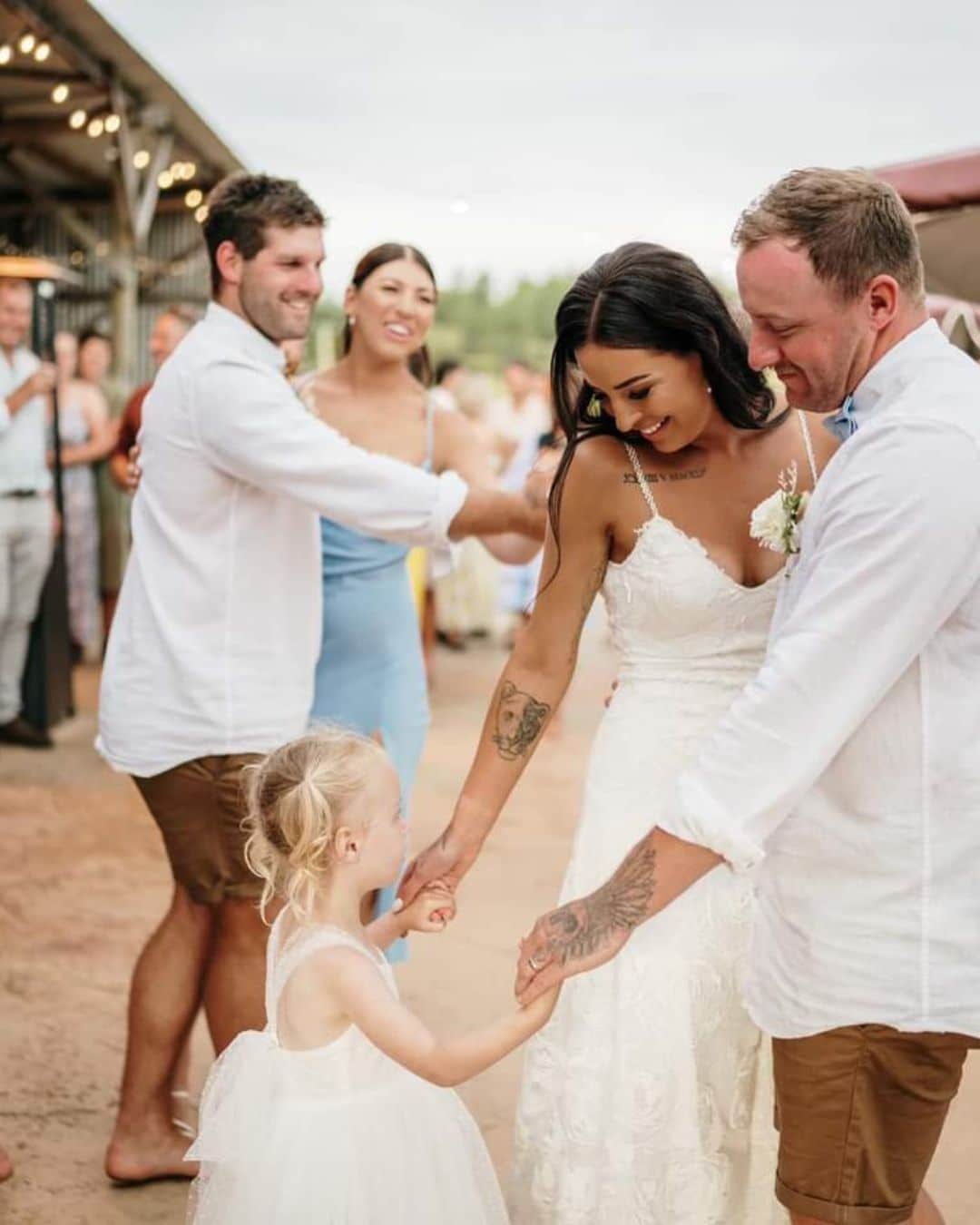 A bride and groom hold hands with a young girl in a white dress at an outdoor wedding, guests visible in the background.