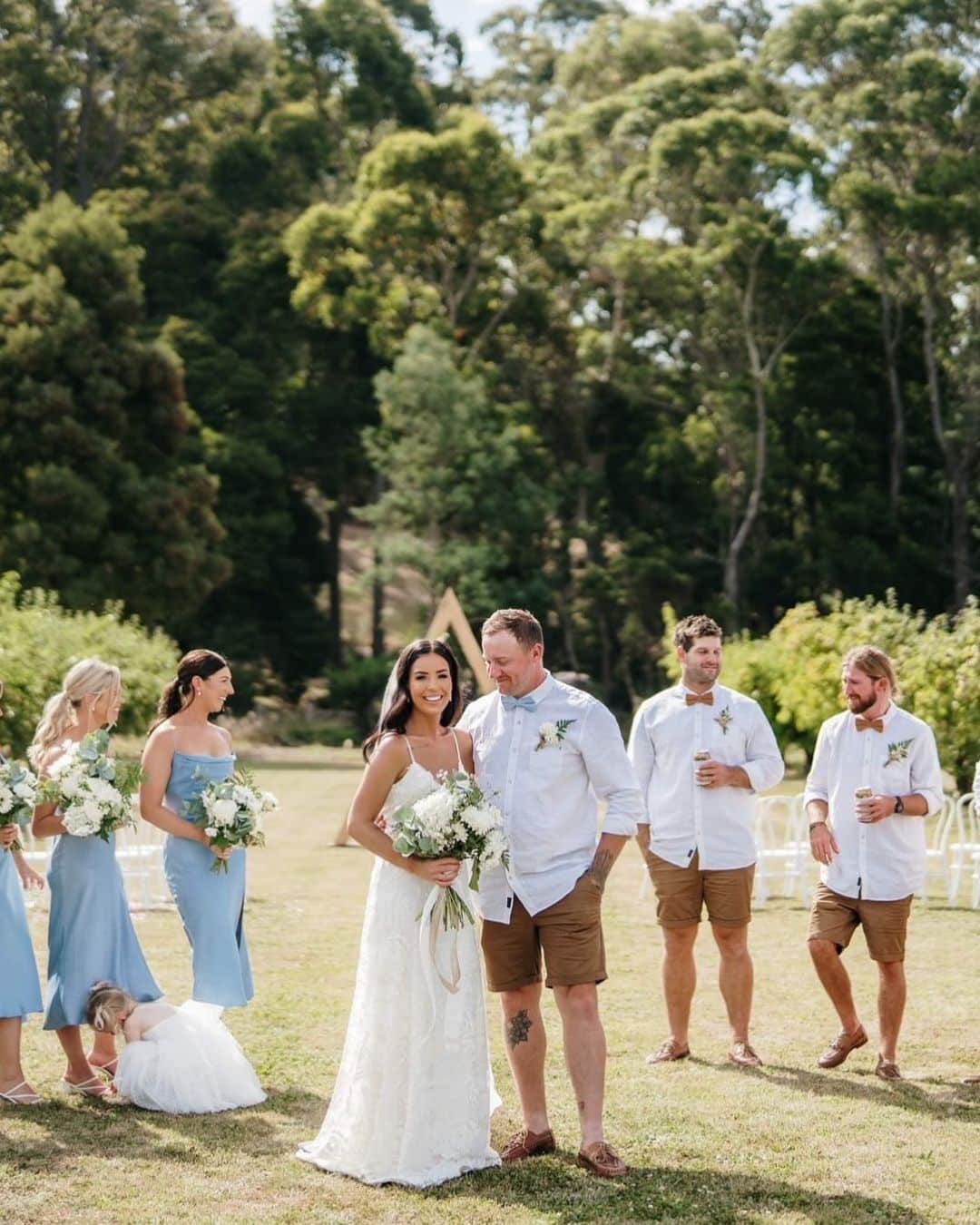 Bride in a white dress and groom in white shirt and brown shorts with bridesmaids in blue dresses and similarly-dressed groomsmen outdoors.
