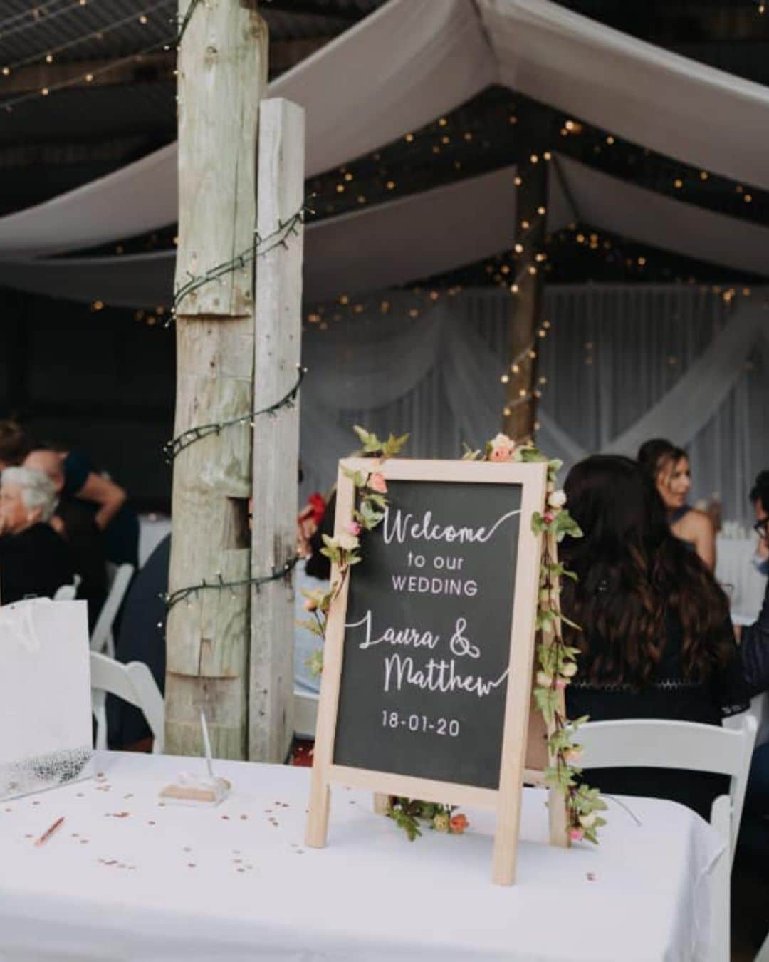 Chalkboard sign reading "Welcome to our wedding Laura & Matthew 18-01-20" on table; guests seated under draped fabric and fairy lights.