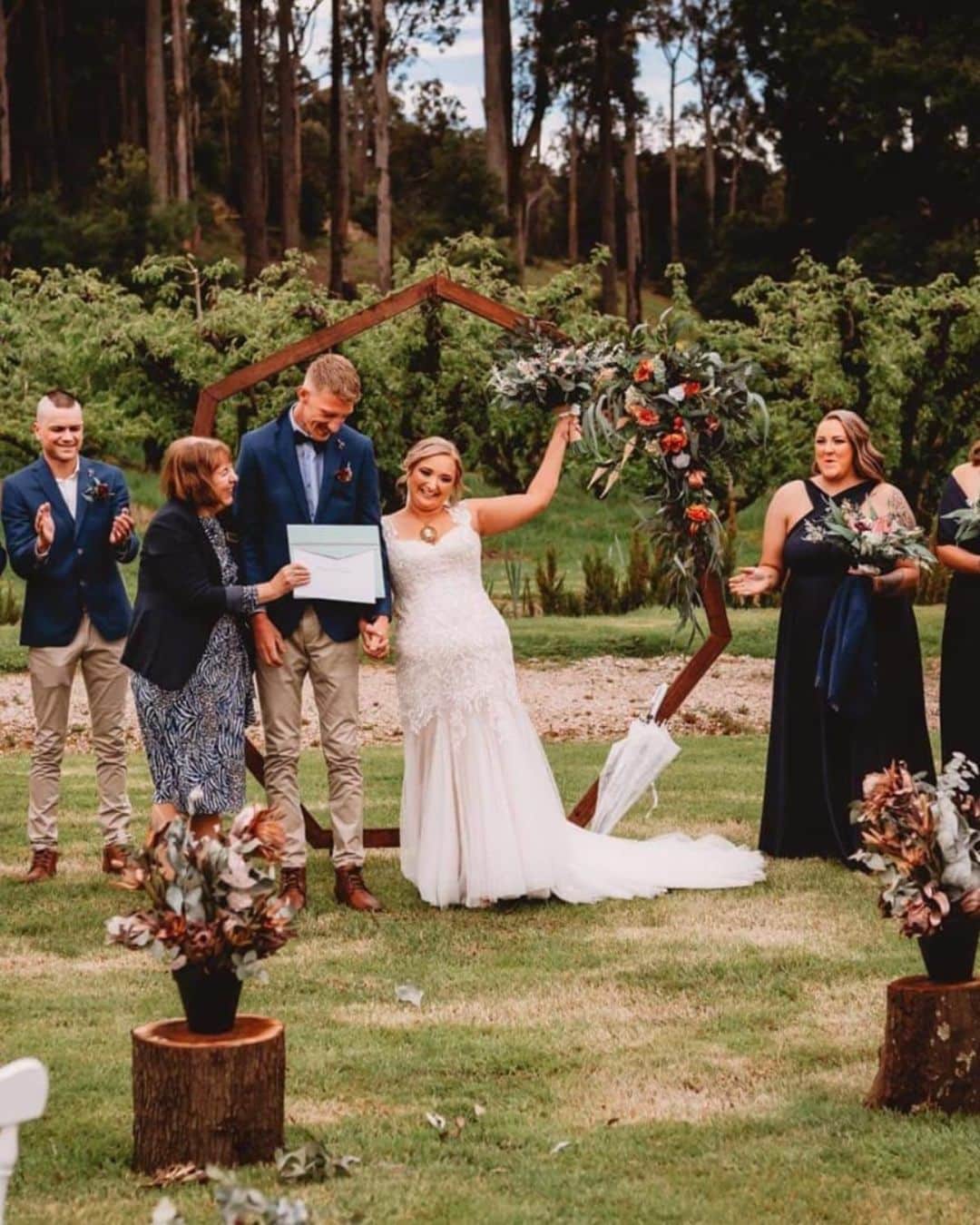 Bride and groom holding a certificate outdoors with wedding party nearby, surrounded by floral arrangements.