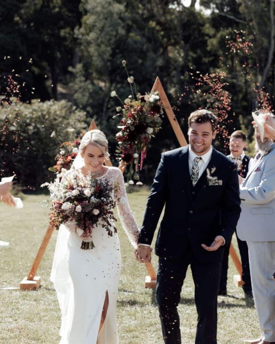 Bride and groom walk hand in hand outdoors, smiling, with confetti falling; floral arrangements and wooden arches behind them.