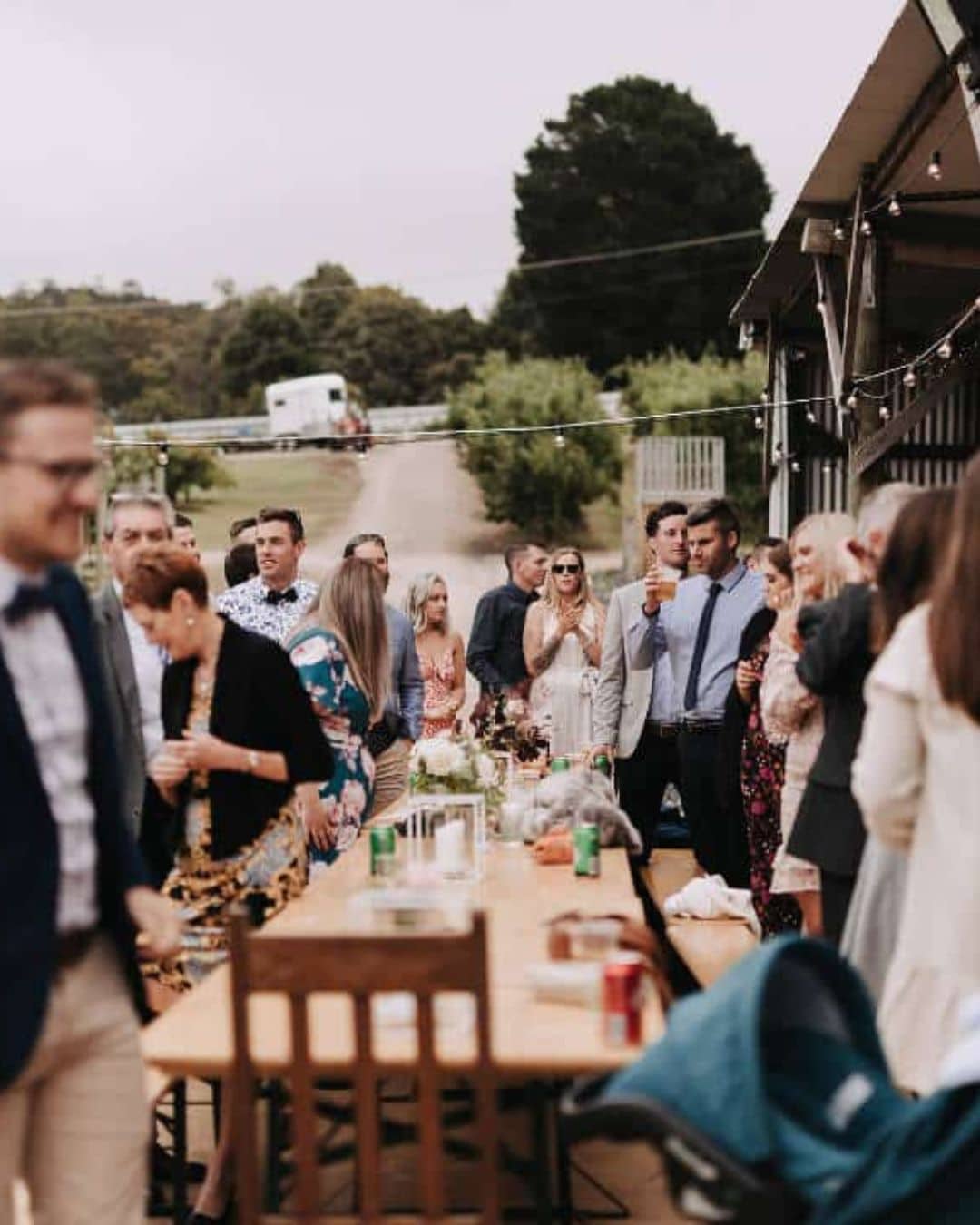 Several people sit and stand around a long outdoor table with drinks and flowers during the day.