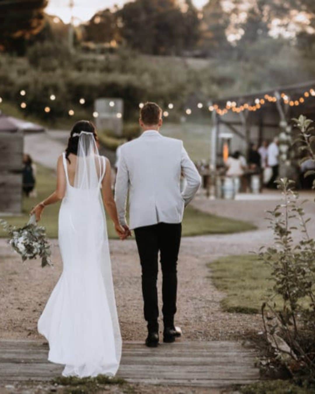 Bride in white dress and veil walks hand in hand with groom outdoors towards reception area with fairy lights.