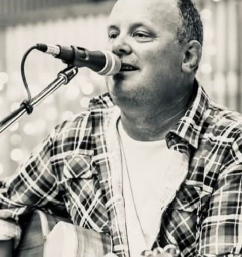Man wearing a checked shirt sings into a microphone and plays an acoustic guitar; black and white image.