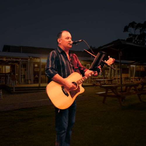 A man playing an acoustic guitar and singing into a microphone outside near picnic tables at twilight.