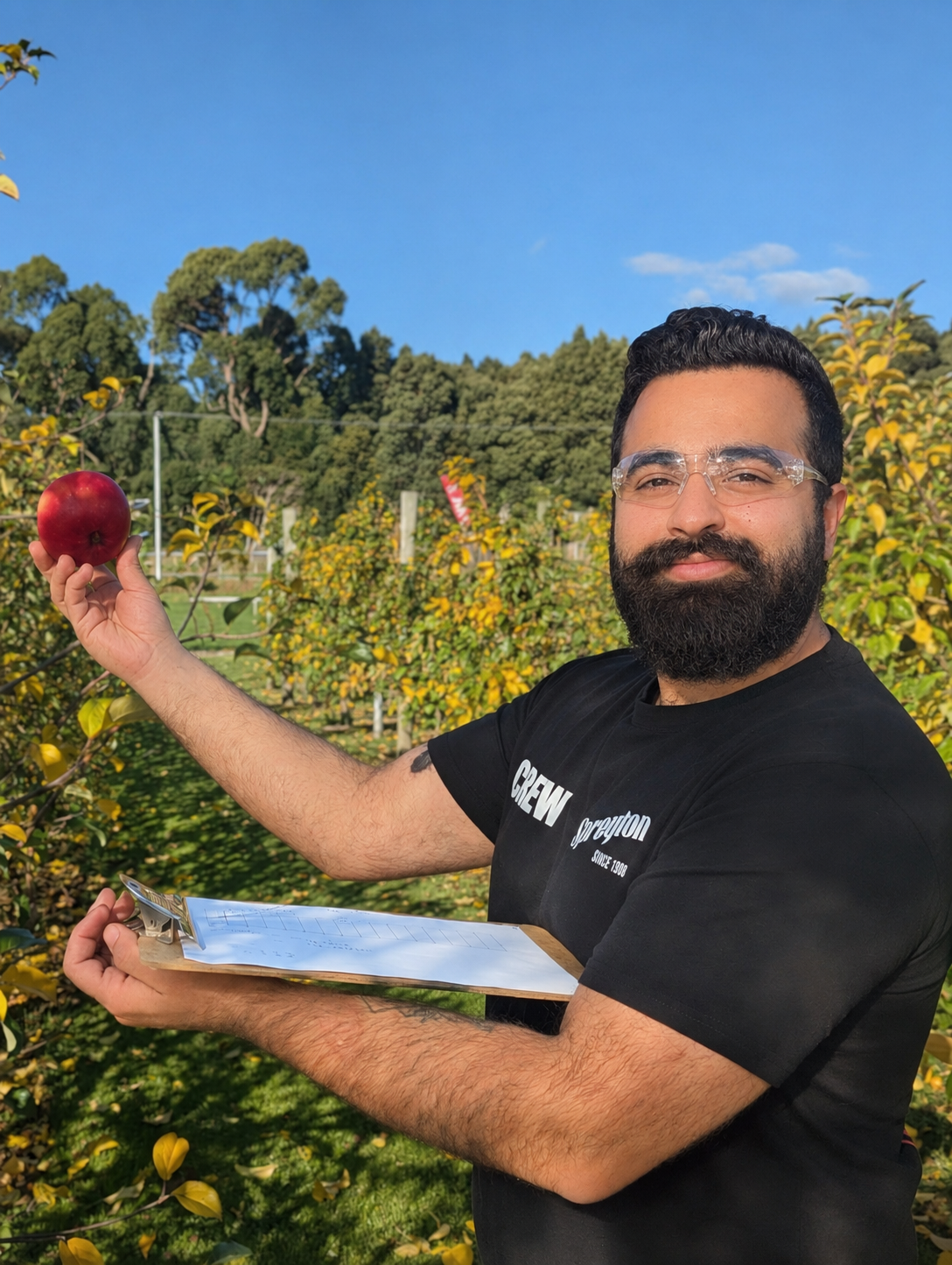 Man in an orchard holding a red apple and a clipboard on a sunny day.