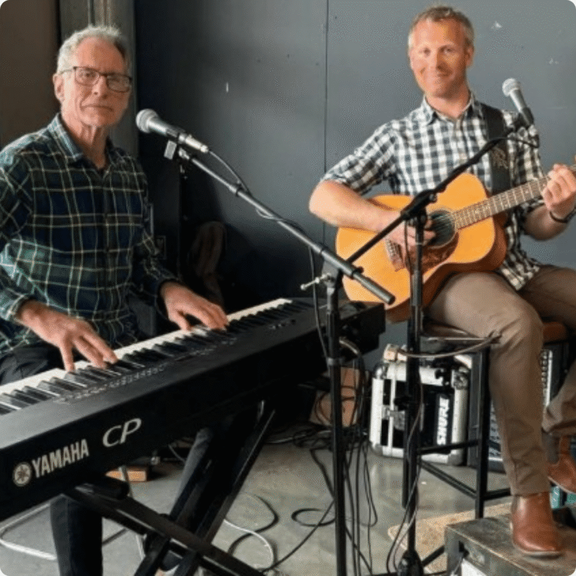 Two men seated indoors, one playing a Yamaha CP keyboard, the other playing an acoustic guitar; both have microphones.