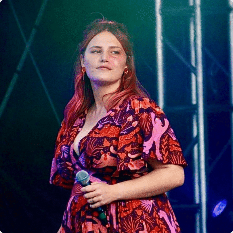 A woman with red hair holds a microphone on stage, wearing a patterned red and purple dress; stage lighting and metal trusses behind.