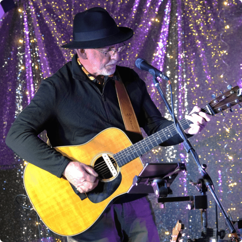 An older man in a black hat and shirt plays an acoustic guitar and sings into a microphone on stage with a sparkly backdrop.