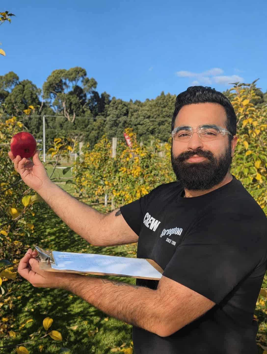Man holding a red apple and clipboard standing in an orchard with trees and a blue sky in the background.