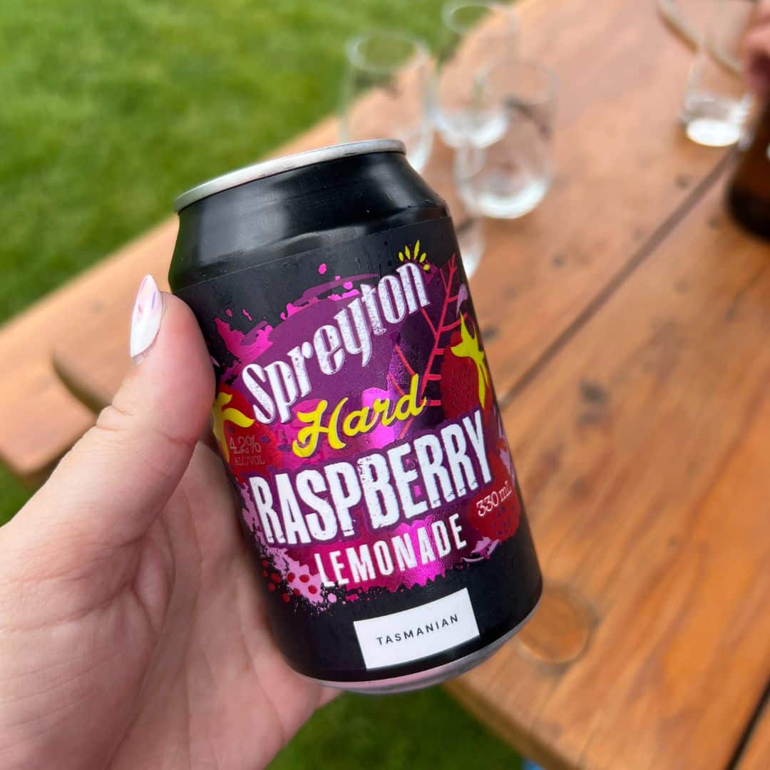 A hand holds a Spreyton Hard Raspberry Lemonade tin outdoors; wooden table and clear glasses are in the background.