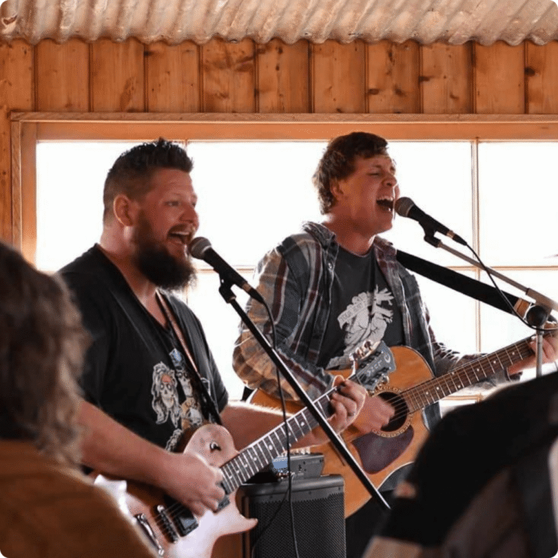 Two men indoors play guitars and sing into microphones, with sunlight streaming through a window behind them.