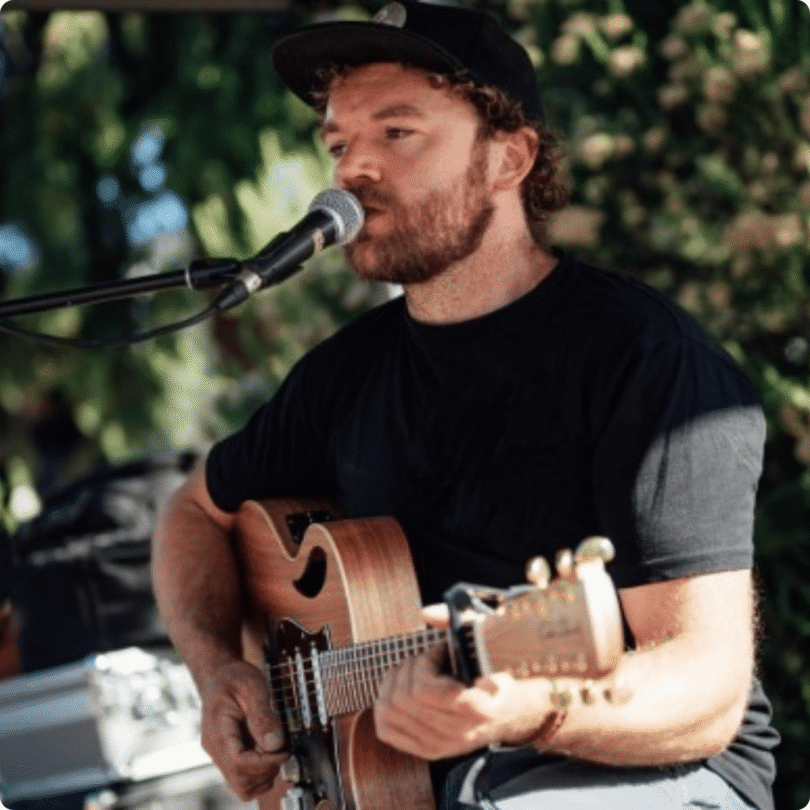 Man in black T-shirt and cap plays acoustic guitar and sings into microphone outdoors.