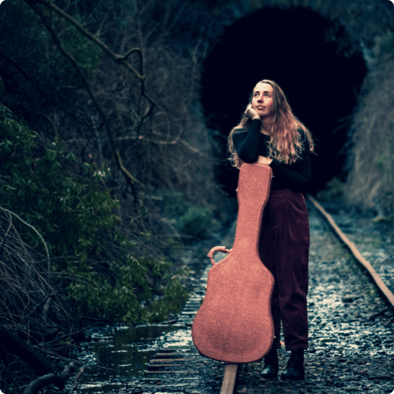 A woman with long hair stands on railway tracks near a tunnel, leaning on a guitar case.