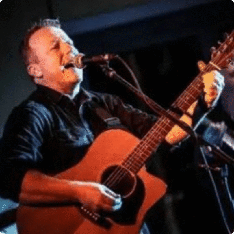 A man playing an acoustic guitar and singing into a microphone on a stage with dramatic lighting.
