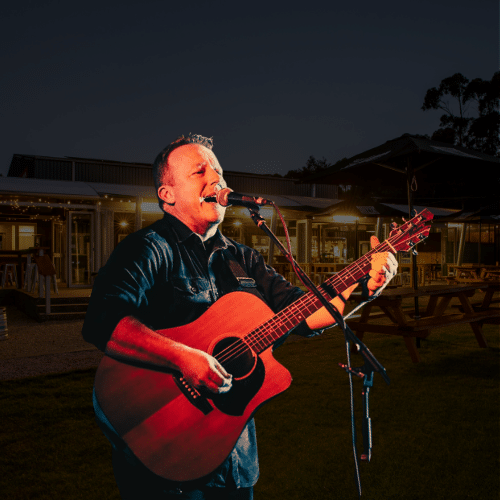 A man plays an acoustic guitar and sings into a microphone outdoors at night in front of a lit building and picnic tables.