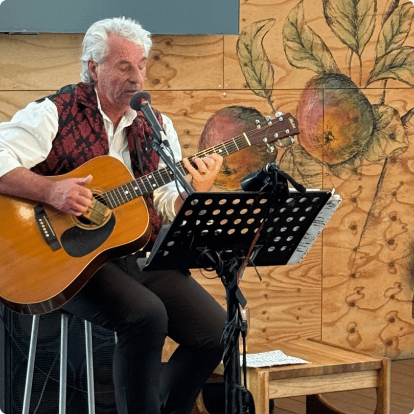 An older man sits, playing an acoustic guitar and singing into a microphone, with a music stand and fruit art behind him.
