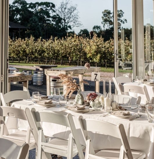 Tables covered with white cloth and surrounded by chairs are arranged in a glass-walled space, with views of a vineyard and outdoor seating area on a sunny day.
