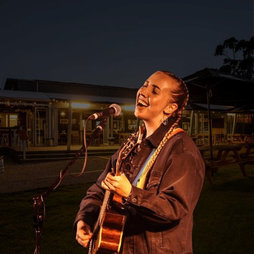 A woman plays acoustic guitar and sings into a microphone outdoors at night, with lit buildings and picnic tables in the background.