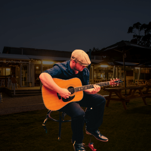 A man wearing a cap sits on a stool and plays an acoustic guitar outdoors at night, with buildings and picnic tables in the background.