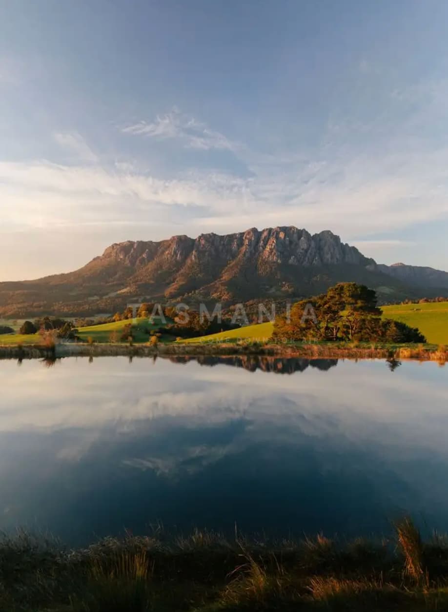 Mountain range reflected in a calm lake, grassy fields and trees in the foreground, partly cloudy sky.